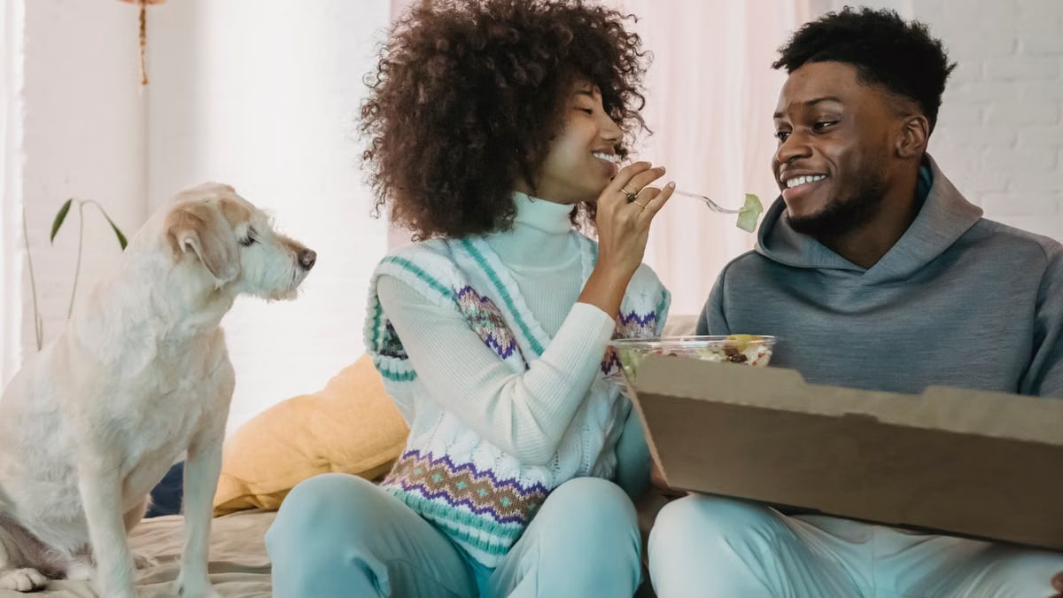 Happy African American couple eating fresh organic salad together while cute white domestic dog watching